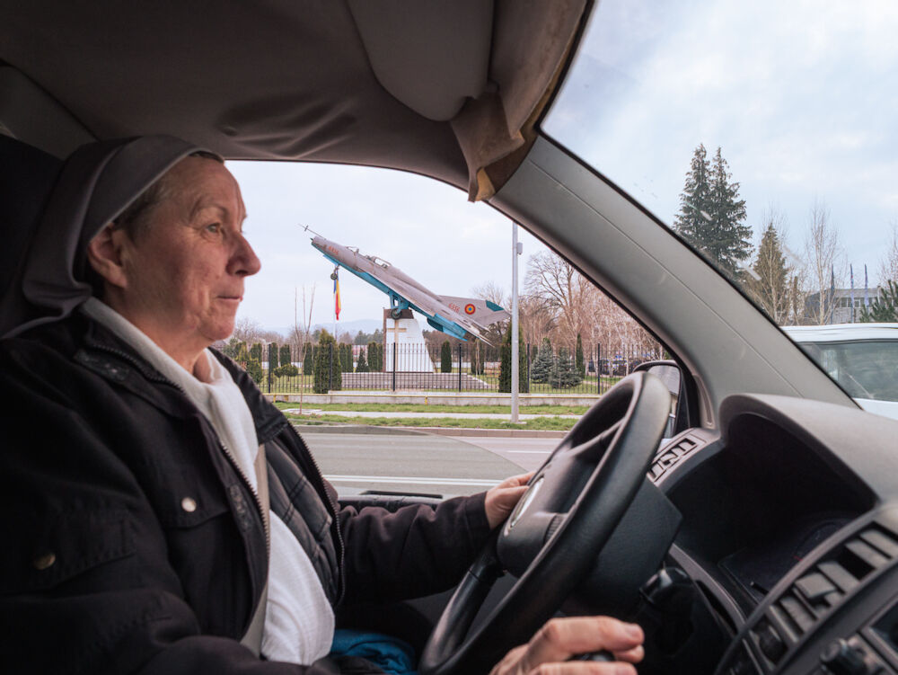 Sœur Monica pendant un déplacement entre deux familles dans la ville de Bacău. En arrière-plan, un chasseur-bombardier soviétique transformé en monument aux pilotes de la ville, dressé dans l'enceinte d'une église.